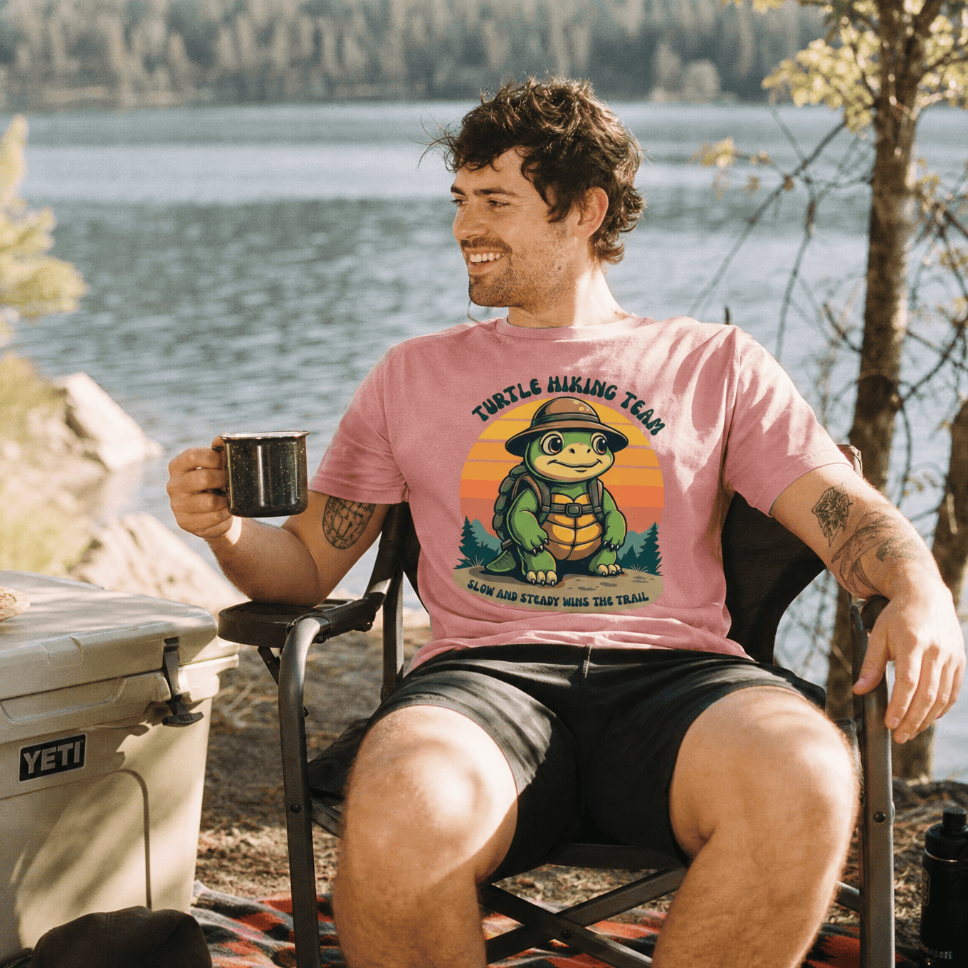 A man sits by a lake wearing a pink graphic t-shirt featuring a turtle and the words "Turtle Hiking Team" along with "Slow and Steady Wins the Trail.