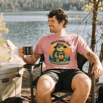 A man sits by a lake wearing a pink graphic t-shirt featuring a turtle and the words "Turtle Hiking Team" along with "Slow and Steady Wins the Trail.