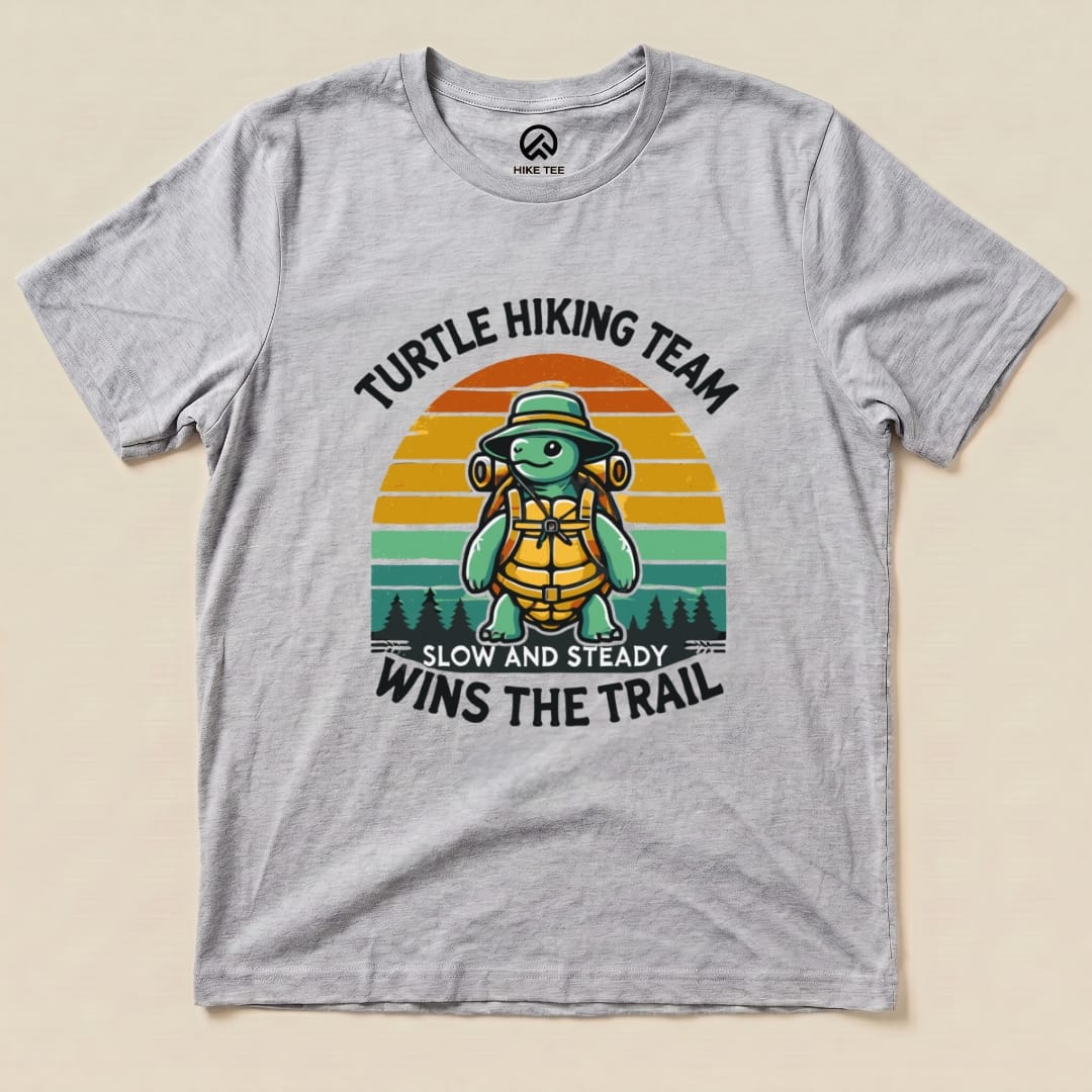 A graphic t-shirt featuring a cheerful turtle in hiking gear, with the phrase "Turtle Wins the Trail" and colorful sunset background.