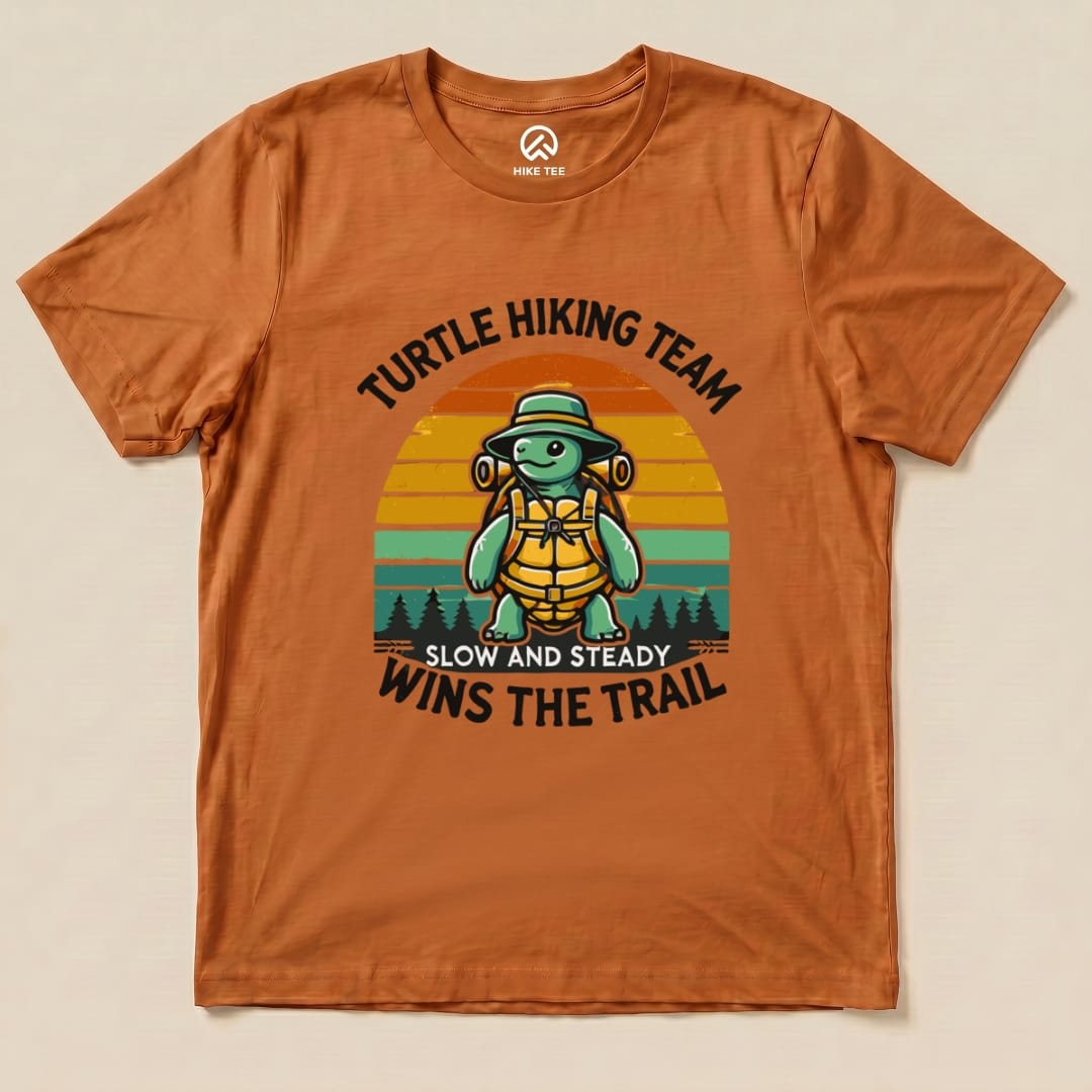 Graphic t-shirt in orange featuring a turtle with a hiking backpack and the slogan "Turtle Wins the Trail" above a sunset background.