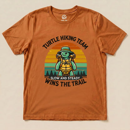 Graphic t-shirt in orange featuring a turtle with a hiking backpack and the slogan "Turtle Wins the Trail" above a sunset background.