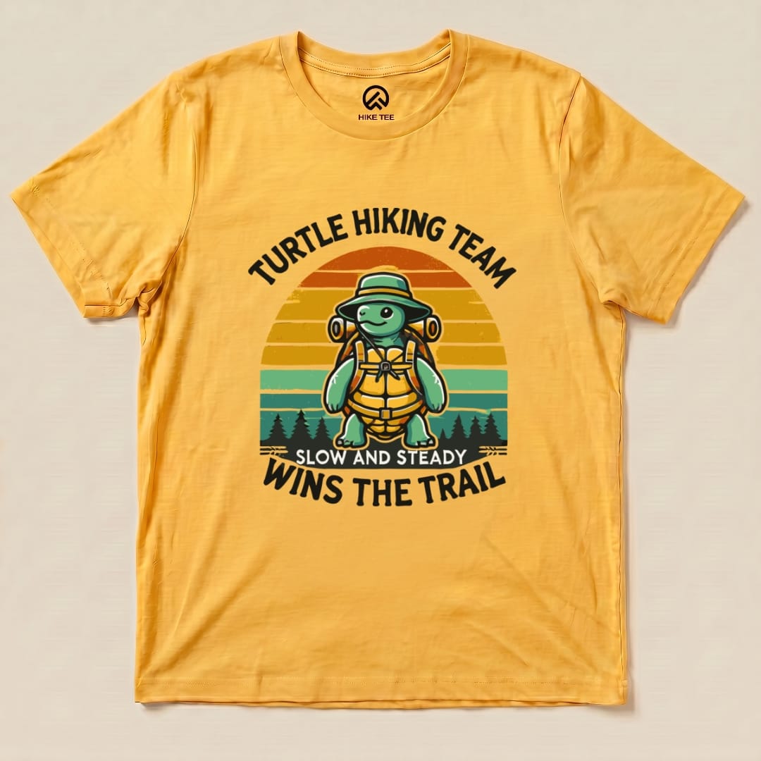 A flat graphic t-shirt in yellow showcases a turtle wearing a hat and backpack with the text "Turtle Wins the Trail".