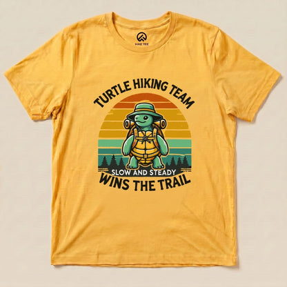 A flat graphic t-shirt in yellow showcases a turtle wearing a hat and backpack with the text "Turtle Wins the Trail".