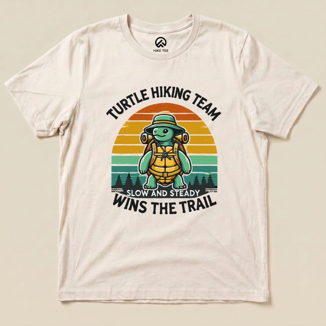 A cream graphic t-shirt featuring a turtle in a hiking hat and backpack, with the phrase "Turtle Wins the Trail" printed in bold.