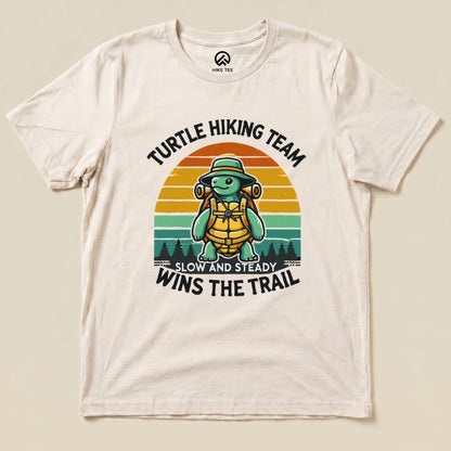 A cream graphic t-shirt featuring a turtle in a hiking hat and backpack, with the phrase "Turtle Wins the Trail" printed in bold.