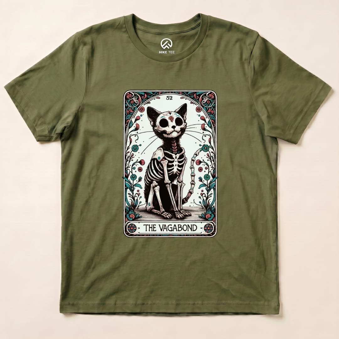 A graphic t-shirt in olive green featuring a whimsical skeleton cat design, titled "The Vagabond," surrounded by decorative elements.