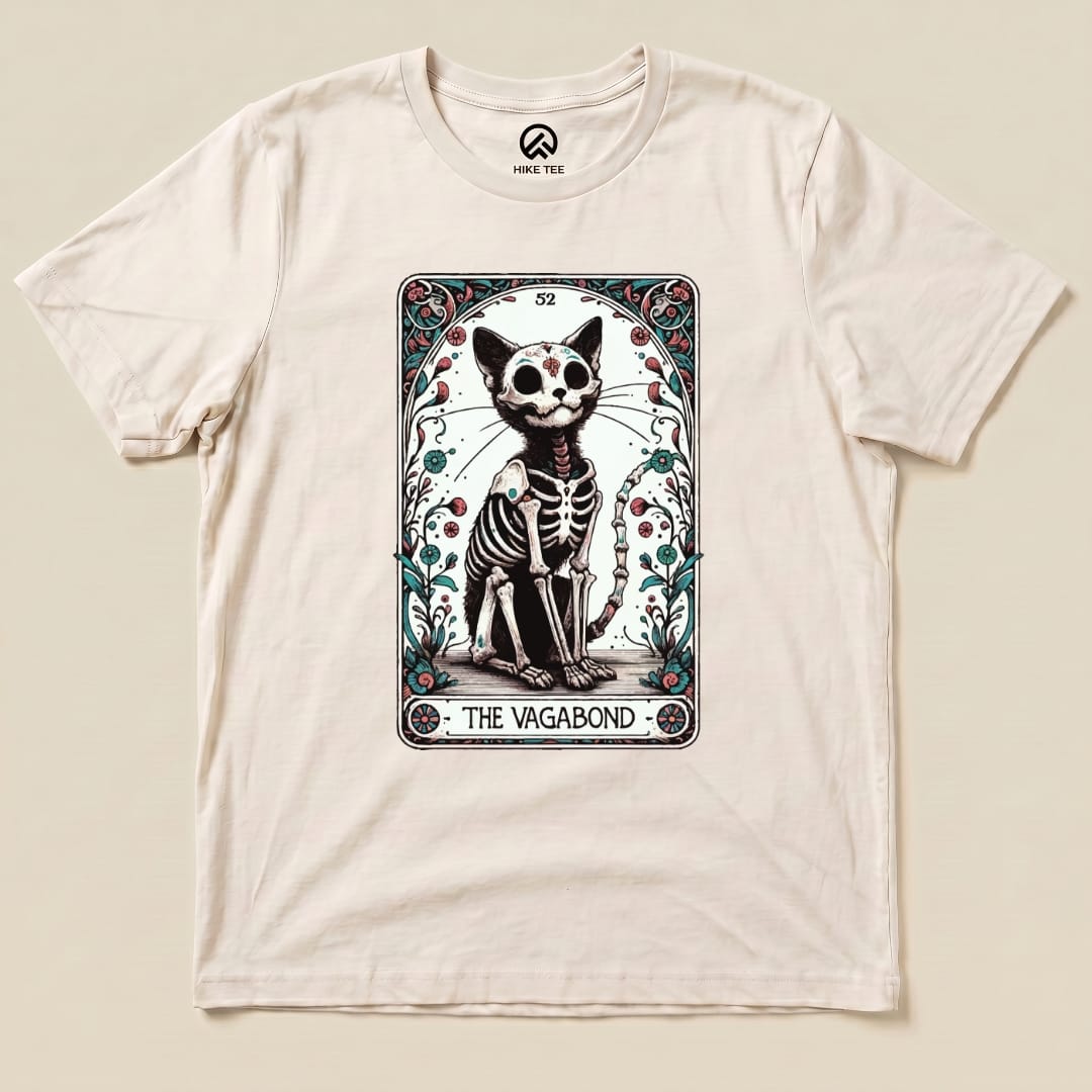 A graphic t-shirt featuring a cartoonish skeleton cat design inside a tarot card frame, labeled "THE VAGABOND.