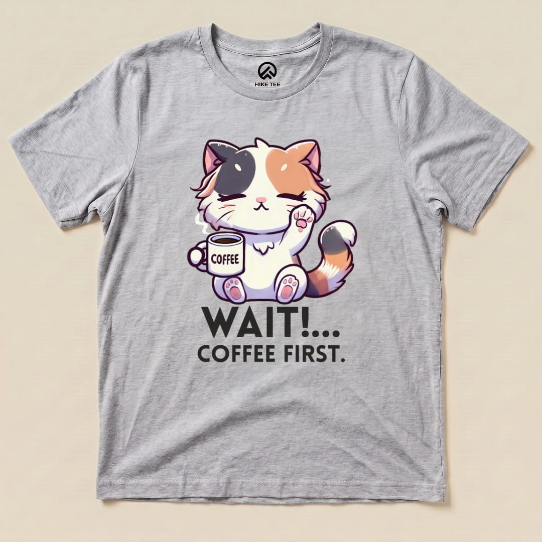 A graphic t-shirt featuring a cute cat holding a coffee cup with the text "WAIT!... COFFEE FIRST" printed below it.