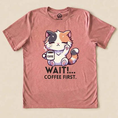 A graphic t-shirt in soft pink featuring a cute cat holding a coffee cup with the slogan "WAIT!... COFFEE FIRST." printed below.