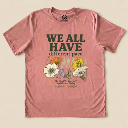 A graphic t-shirt in a soft pink hue featuring the slogan "WE ALL HAVE different pace" surrounded by colorful flowers.