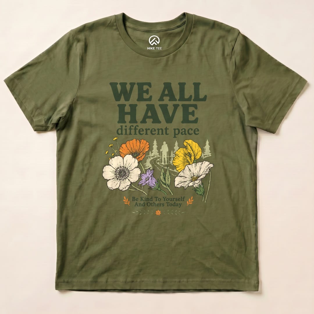 Graphic t-shirt in olive green features the slogan "WE ALL HAVE different pace" along with colorful flowers and a hiker design.