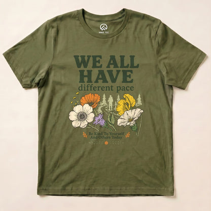 Graphic t-shirt in olive green features the slogan "WE ALL HAVE different pace" along with colorful flowers and a hiker design.