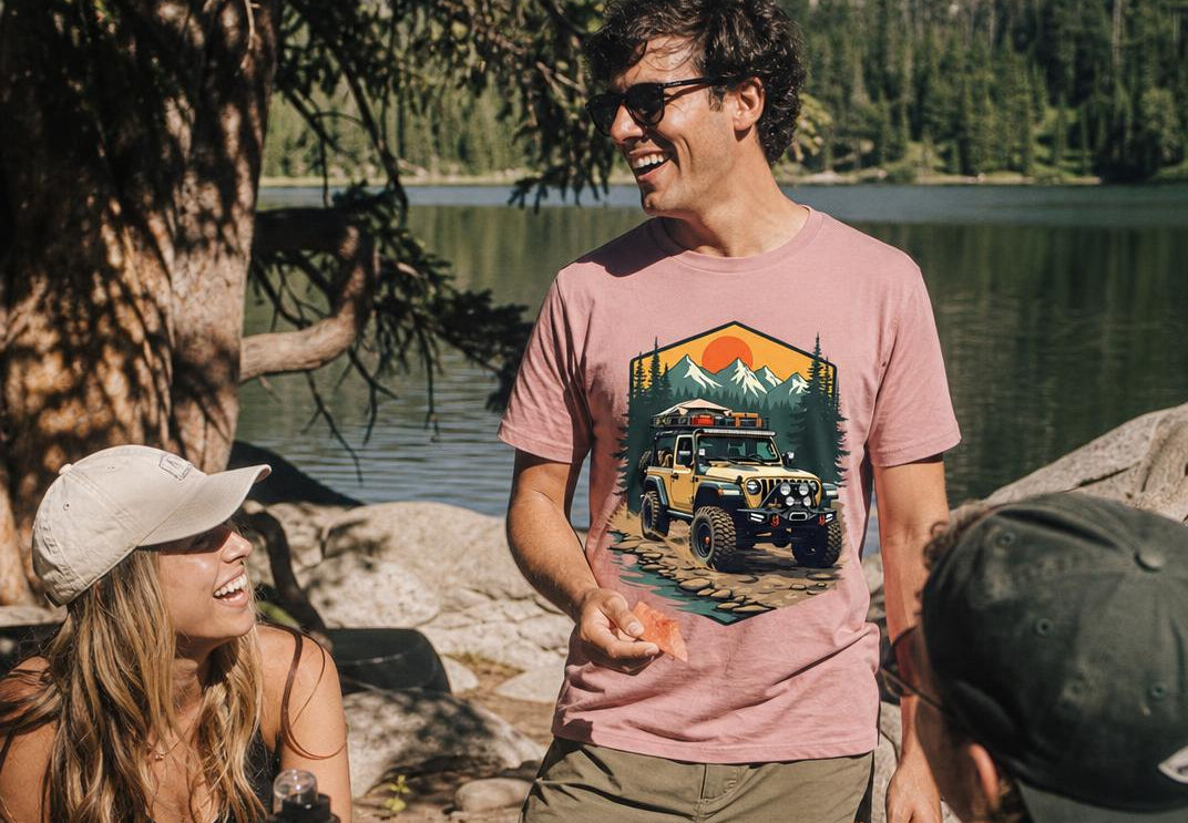 A man wearing a dusty rose pink t-shirt with a graphic of a Jeep and mountains, standing by a lake at a picnic table with a woman and another person.