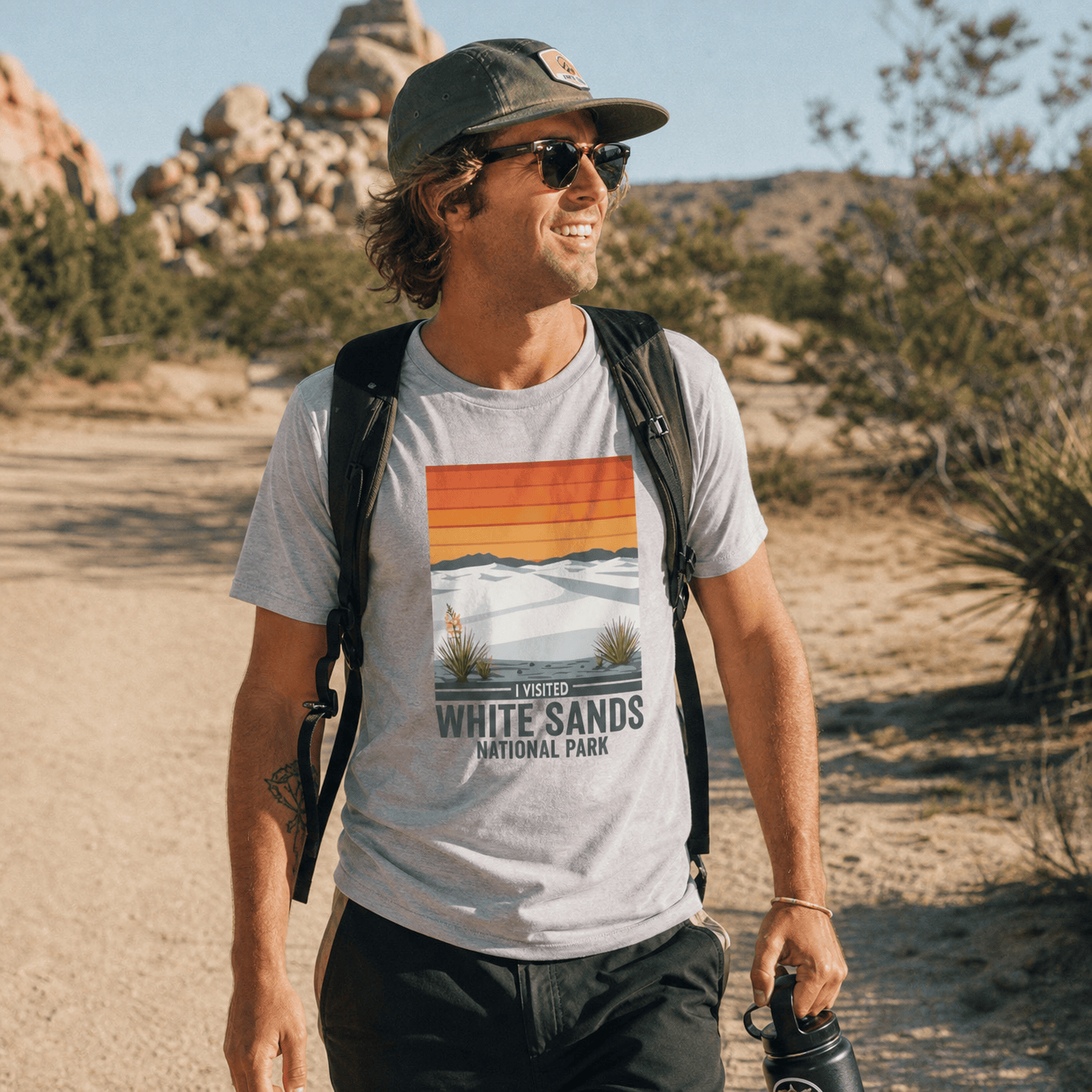 A smiling man hikes on a trail wearing a graphic t-shirt featuring the phrase "I Visited White Sands National Park.