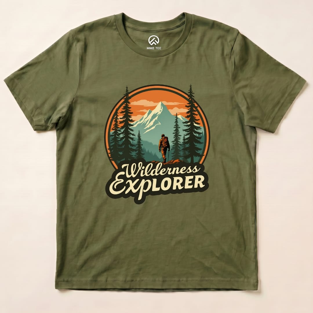 A flat olive green graphic t-shirt features a design of a hiker in front of mountains and trees with the text "Wilderness Explorer Shirt.