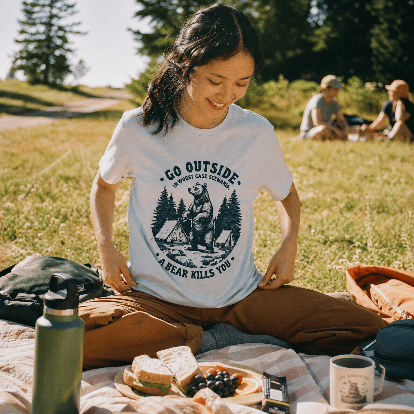 A woman sits on a picnic blanket in a park, wearing a graphic t-shirt that says "Go Outside. In Worst Case Scenario, A Bear Kills You.