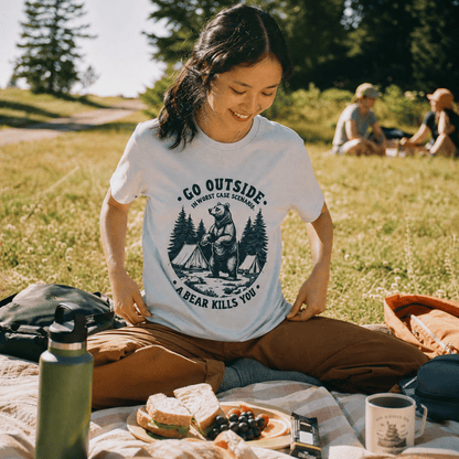 A woman sits on a picnic blanket in a park, wearing a graphic t-shirt that says "Go Outside. In Worst Case Scenario, A Bear Kills You.
