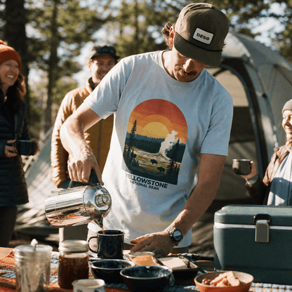 A man pours coffee while wearing a graphic t-shirt featuring a scenic illustration of Yellowstone National Park.