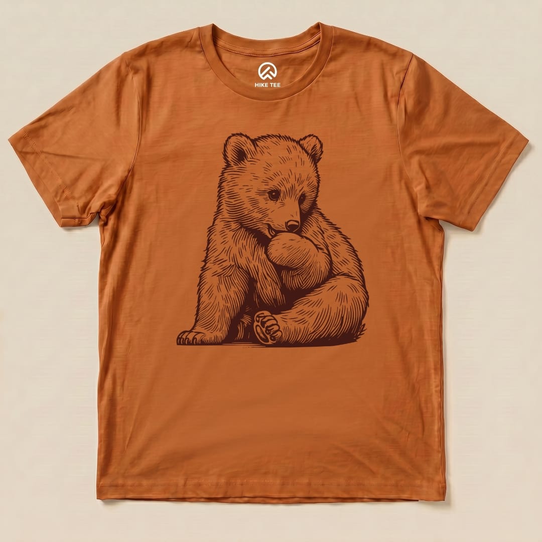 A graphic t-shirt featuring a cute illustration of a bear sitting and nibbling on its paw, the Youth Bear Shirt is in a warm orange hue.