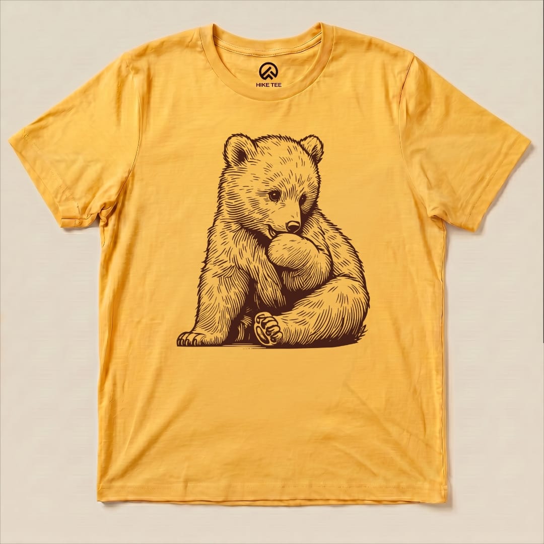 A yellow graphic t-shirt displays an illustration of a cute bear sitting and holding its foot, labeled as a Youth Bear Shirt.