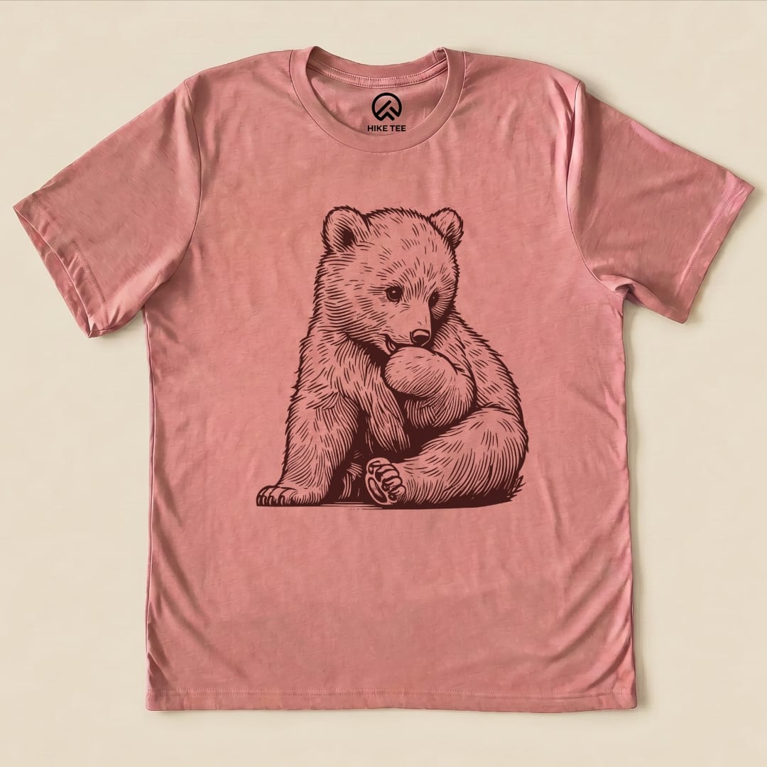A pink graphic t-shirt featuring a cute bear cub sitting and playing with a paw, labeled as the "Youth Bear Shirt.