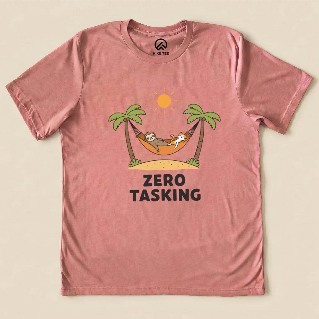 A pink graphic t-shirt featuring a sloth and a cat lounging in a hammock under palm trees, with the phrase "ZERO TASKING.
